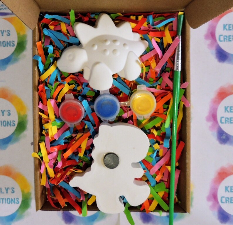 May include: A craft kit with two white ceramic dinosaur figurines, paint pots in red, blue, and yellow, and a green paintbrush. The kit is packaged in a cardboard box with colorful shredded paper.
