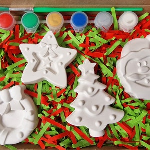 May include: A Christmas-themed paint-your-own ceramic craft kit with five white ceramic ornaments: a star, a Christmas tree, a wreath, a snowman, and a Santa Claus. The kit includes five paint pots in red, green, yellow, blue, and white, and a paintbrush.
