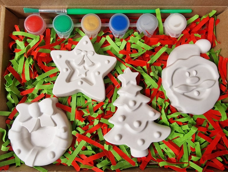 May include: A Christmas-themed paint-your-own ceramic craft kit with five white ceramic ornaments: a star, a Christmas tree, a wreath, a snowman, and a Santa Claus. The kit includes five paint pots in red, green, yellow, blue, and white, and a paintbrush.