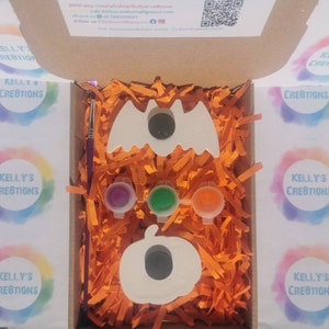 May include: A Halloween-themed craft kit in an open cardboard box. The kit includes a white bat and pumpkin, three paint pots, a paintbrush, and orange paper filler. The box has the text "KELLY'S CREATIONS" on the side.
