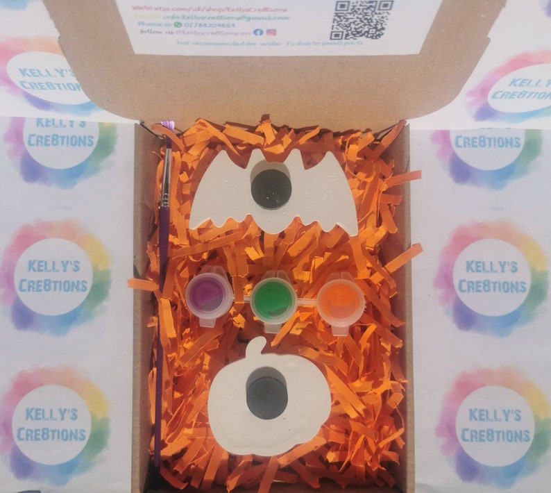 May include: A Halloween-themed craft kit in an open cardboard box. The kit includes a white bat and pumpkin, three paint pots, a paintbrush, and orange paper filler. The box has the text "KELLY'S CREATIONS" on the side.