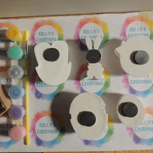 May include: Five white ceramic ornaments with magnets on the back. The ornaments are shaped like a bunny, a tooth, a cloud, a circle, and a crescent moon. The ornaments are surrounded by colorful paint and paintbrushes.