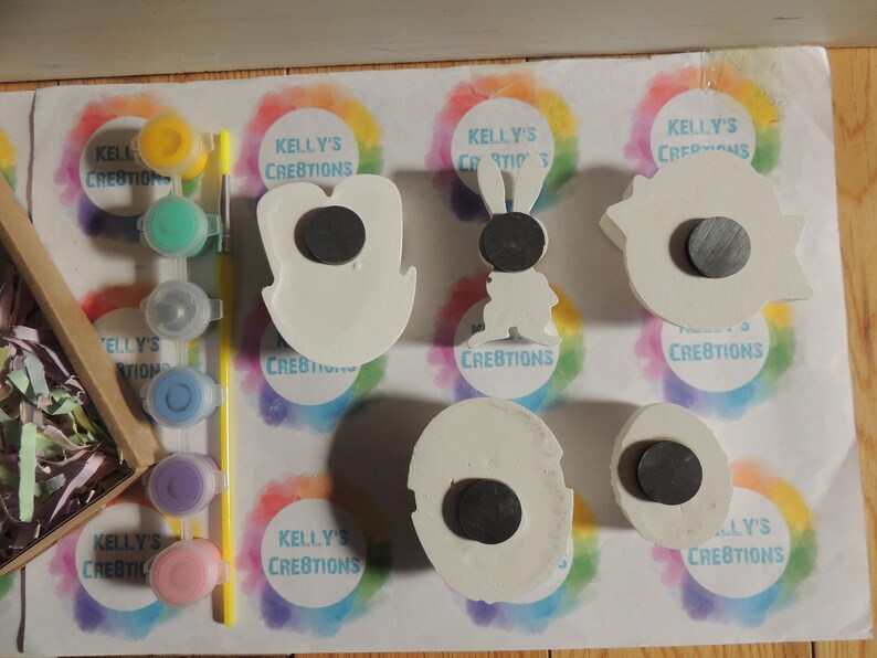 May include: Five white ceramic ornaments with magnets on the back. The ornaments are shaped like a bunny, a tooth, a cloud, a circle, and a crescent moon. The ornaments are surrounded by colorful paint and paintbrushes.