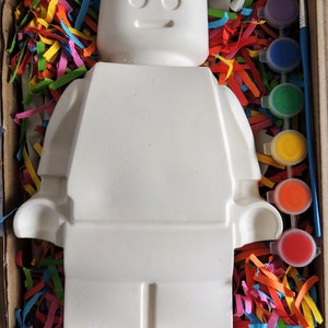 May include: A white ceramic figurine of a Lego person, with a paint-by-numbers design, in a box with colorful paper shreds and a set of paint pots.