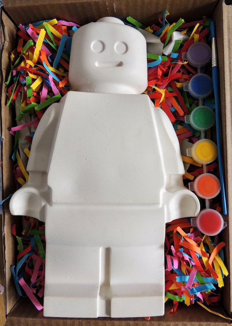 May include: A white ceramic figurine of a Lego person, with a paint-by-numbers design, in a box with colorful paper shreds and a set of paint pots.