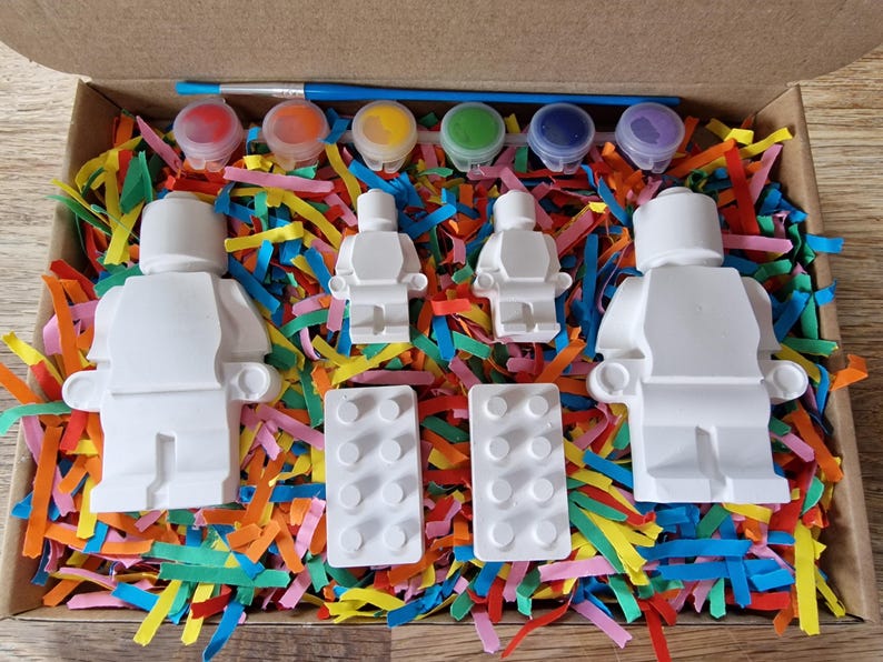 May include: A paint-your-own Lego minifigure set with four white figures, two white Lego baseplates, and six small paint pots with red, orange, yellow, green, blue, and purple paint.