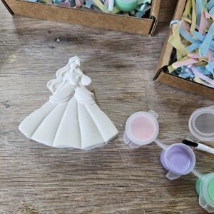 Op de afbeelding: Een wit gipsen beeldje van een prinses in een lange jurk met golvend haar, vergezeld van een klein penseel en verschillende kleine plastic potjes met pastelkleurige verf, waaronder roze, paarse en lichtgroene tinten.