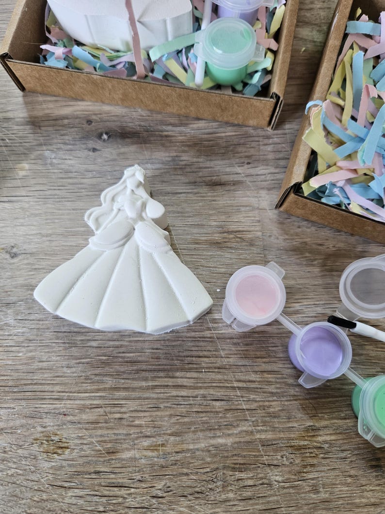 Op de afbeelding: Een wit gipsen beeldje van een prinses in een lange jurk met golvend haar, vergezeld van een klein penseel en verschillende kleine plastic potjes met pastelkleurige verf, waaronder roze, paarse en lichtgroene tinten.