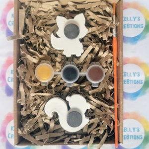 May include: A craft kit with two white ceramic animal figurines, a squirrel and a fox, ready to be painted. The kit includes three small paint pots, one yellow, one black, and one brown, and a paintbrush.