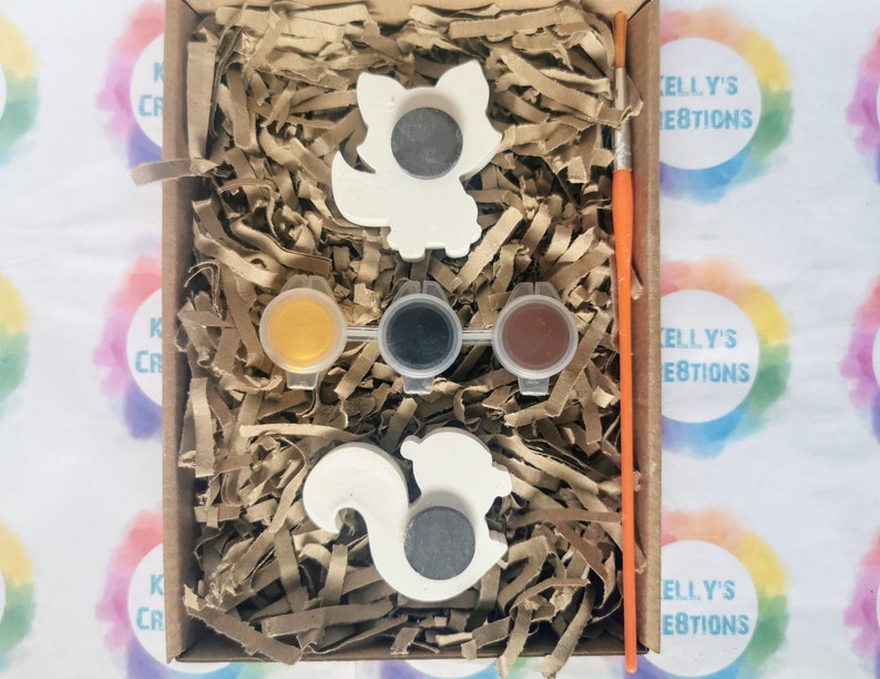 May include: A craft kit with two white ceramic animal figurines, a squirrel and a fox, ready to be painted. The kit includes three small paint pots, one yellow, one black, and one brown, and a paintbrush.