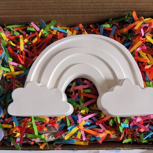 May include: A white ceramic rainbow with clouds on either side, sitting in a box filled with colorful shredded paper. A set of paint pots and a paintbrush are included.