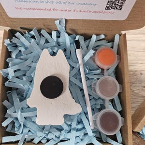 May include: A craft kit in an open cardboard box, with a white ceramic figure with a black magnet, paint pots, and a paintbrush. The box contains blue paper shreds. Text on the box reads: Open paints and use brush to start creating.
