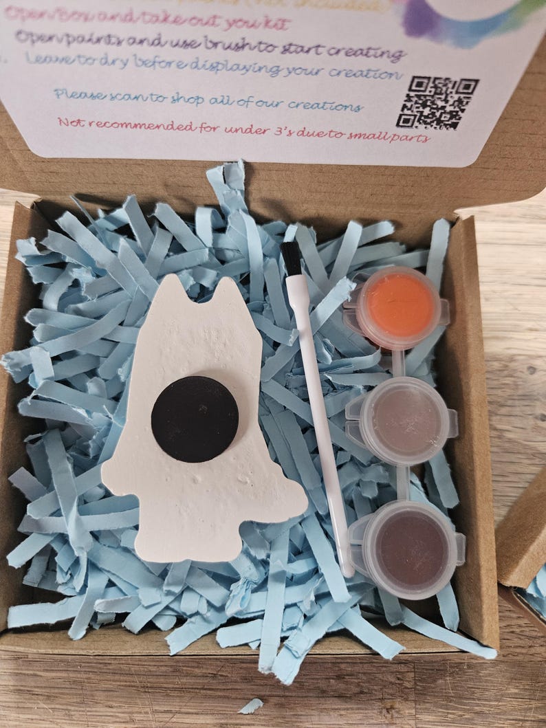 May include: A craft kit in an open cardboard box, with a white ceramic figure with a black magnet, paint pots, and a paintbrush. The box contains blue paper shreds. Text on the box reads: Open paints and use brush to start creating.