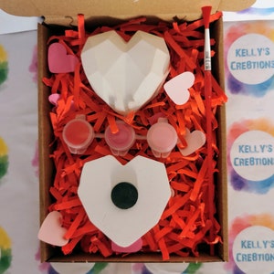 May include: A craft kit with two white ceramic heart-shaped ornaments, three small pots of paint, a paintbrush, and pink paper hearts in a cardboard box filled with red shredded paper. The box has a label that says "Kelly's Creations".