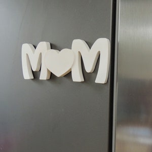 Pu&ograve; includere: Lettere in legno bianco che compongono la parola "MOM" con un cuore bianco tra le "M". Le lettere sono attaccate a un frigorifero grigio.