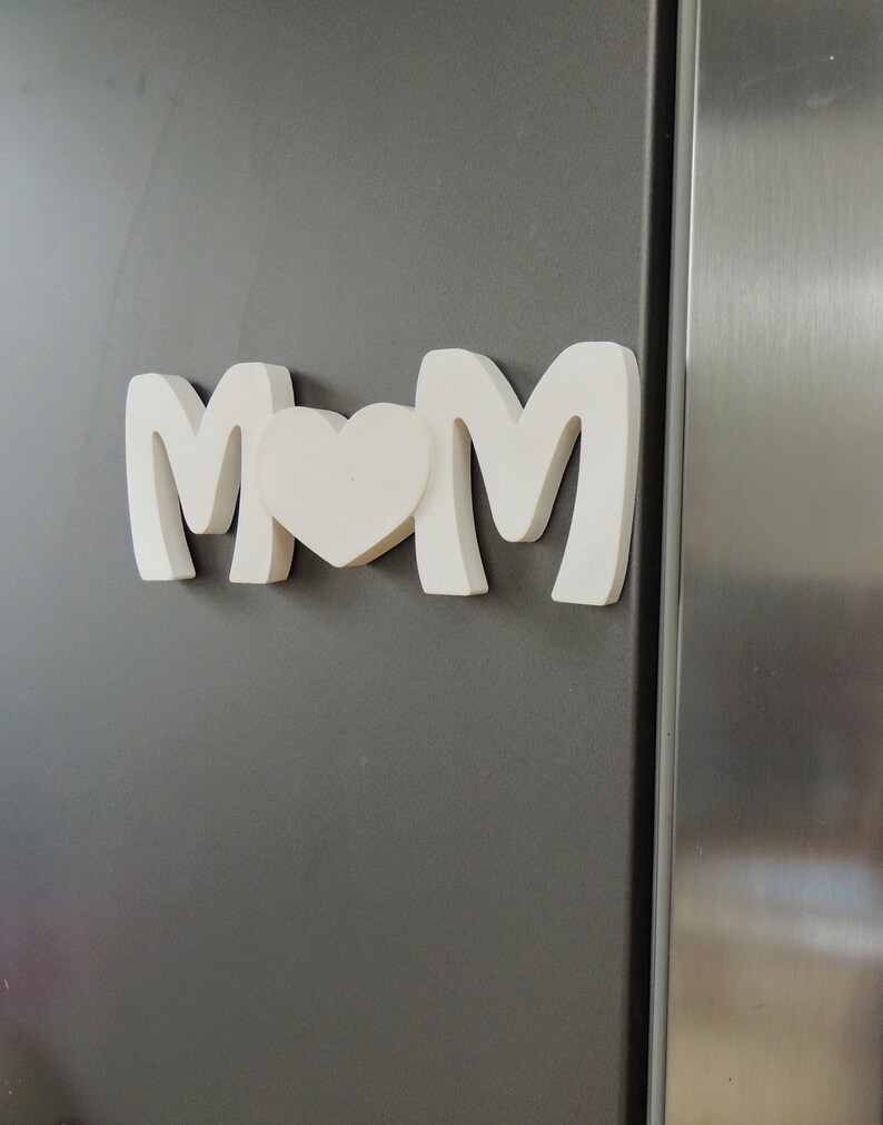 Pu&ograve; includere: Lettere in legno bianco che compongono la parola "MOM" con un cuore bianco tra le "M". Le lettere sono attaccate a un frigorifero grigio.