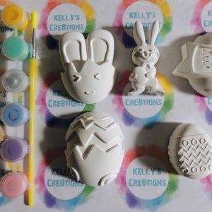 May include: A set of six white ceramic Easter-themed ornaments, including a bunny, an egg with a chevron pattern, an egg with a circle pattern, an egg with a zig-zag pattern, a bunny with ears, and a banner that reads "Happy Easter". The ornaments are ready to be painted.