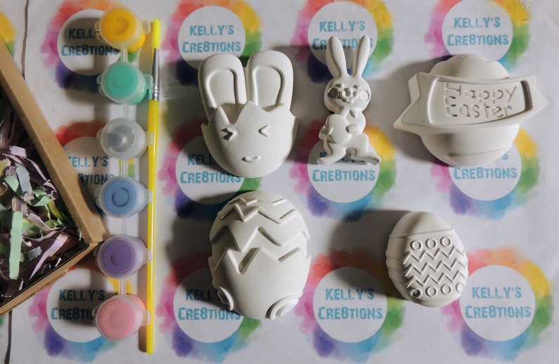 May include: A set of six white ceramic Easter-themed ornaments, including a bunny, an egg with a chevron pattern, an egg with a circle pattern, an egg with a zig-zag pattern, a bunny with ears, and a banner that reads "Happy Easter". The ornaments are ready to be painted.