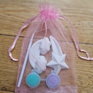 Puede incluir: Una bolsa de organza rosa llena de un arco&iacute;ris, una estrella y una estrella fugaz de cer&aacute;mica blanca, junto con dos peque&ntilde;os botes de pintura, uno turquesa y otro lavanda.