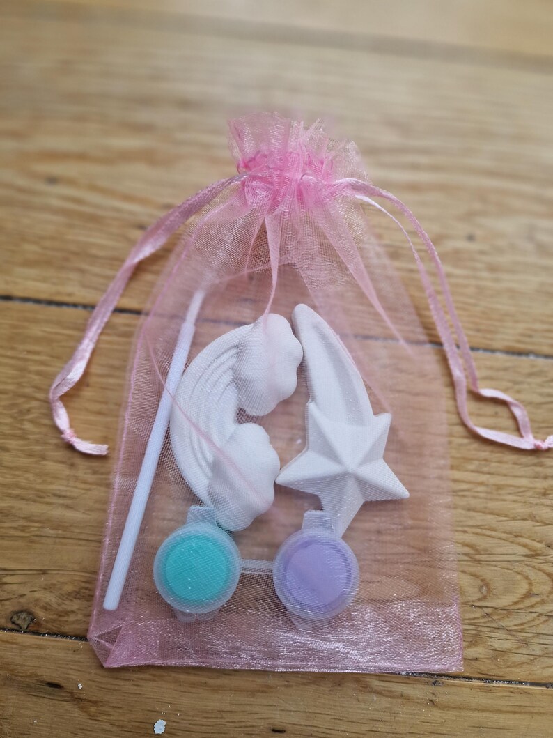Puede incluir: Una bolsa de organza rosa llena de un arco&iacute;ris, una estrella y una estrella fugaz de cer&aacute;mica blanca, junto con dos peque&ntilde;os botes de pintura, uno turquesa y otro lavanda.