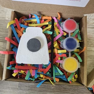 May include: A craft kit in a cardboard box containing a white ceramic shape with a black circular magnet on the back, a thin black paintbrush, and three small plastic pots of red, blue, and yellow paint, all nestled in colorful paper shreds.