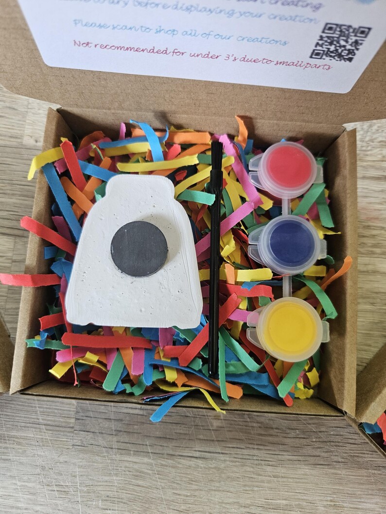 May include: A craft kit in a cardboard box containing a white ceramic shape with a black circular magnet on the back, a thin black paintbrush, and three small plastic pots of red, blue, and yellow paint, all nestled in colorful paper shreds.