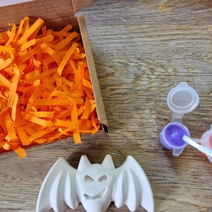 May include: A white ceramic bat figure with outstretched wings, a smiling face, and pointed ears. The bat is next to a small paint set with purple paint and a cardboard box filled with orange paper strips. The items are on a wooden surface.