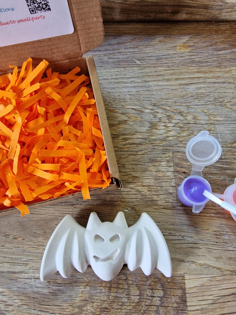 May include: A white ceramic bat figure with outstretched wings, a smiling face, and pointed ears. The bat is next to a small paint set with purple paint and a cardboard box filled with orange paper strips. The items are on a wooden surface.