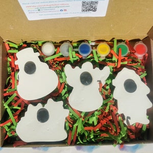 May include: A craft kit in a cardboard box, featuring five white ceramic shapes with black centers, paint pots in various colors, and paintbrushes. The box is filled with red, green, and yellow shredded paper.