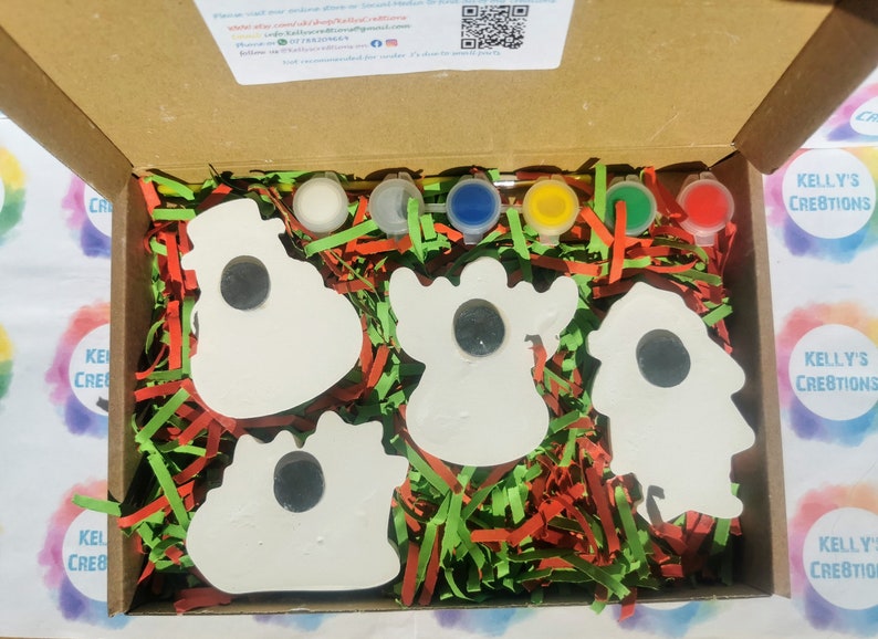 May include: A craft kit in a cardboard box, featuring five white ceramic shapes with black centers, paint pots in various colors, and paintbrushes. The box is filled with red, green, and yellow shredded paper.