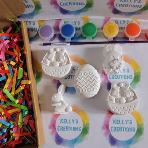 May include: A set of five white ceramic Easter-themed figurines, including a bunny, an egg, and a basket, with paint pots and a brush for decorating. The figurines are on a white background with colorful rainbow patterns.