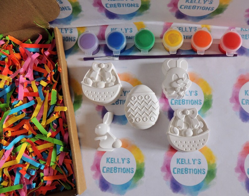 May include: A set of five white ceramic Easter-themed figurines, including a bunny, an egg, and a basket, with paint pots and a brush for decorating. The figurines are on a white background with colorful rainbow patterns.
