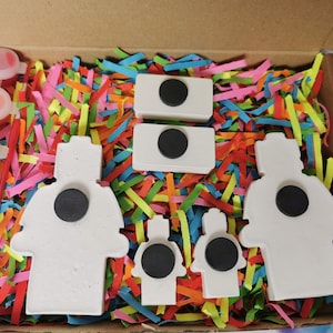 May include: A set of five white ceramic figures with black magnetic circles on the back. The figures are shaped like firefighters and are surrounded by colorful shredded paper. A set of paint pots and a paintbrush are included.