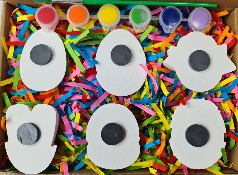 May include: A set of six white ceramic shapes with black magnet circles in the centre, surrounded by colourful shredded paper. The shapes are in a cardboard box with five small paint pots and a paintbrush.