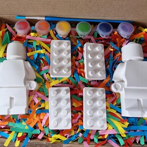 May include: A paint-your-own Lego minifigure kit with two white figurines, four white Lego bricks, and six small paint pots with red, orange, yellow, green, blue, and purple paint. The kit is packaged in a cardboard box with colorful shredded paper.