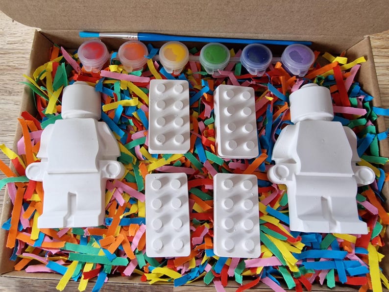 May include: A paint-your-own Lego minifigure kit with two white figurines, four white Lego bricks, and six small paint pots with red, orange, yellow, green, blue, and purple paint. The kit is packaged in a cardboard box with colorful shredded paper.