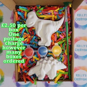 Op de afbeelding: Een knutseldoos met twee witte dinosaurusfiguren, drie verfpotjes en een groene verfkwast. De doos is gevuld met kleurrijk papier. De tekst "KELLY'S CREATION" is zichtbaar op de zijkant van de doos.