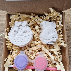 May include: A cardboard box with a white Easter basket and a bunny figurine inside. The box also contains two small pots of paint and a paintbrush. The box has a warning label that says "Not recommended for under 3's due to small parts."