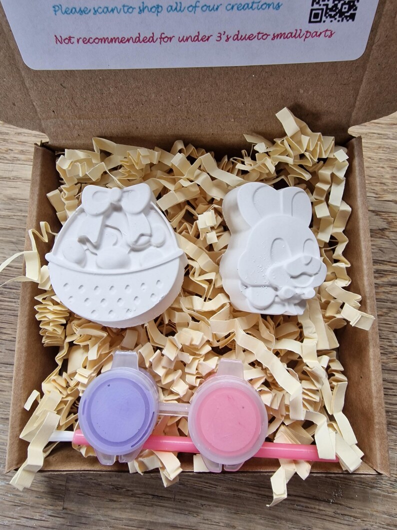 May include: A cardboard box with a white Easter basket and a bunny figurine inside. The box also contains two small pots of paint and a paintbrush. The box has a warning label that says "Not recommended for under 3's due to small parts."