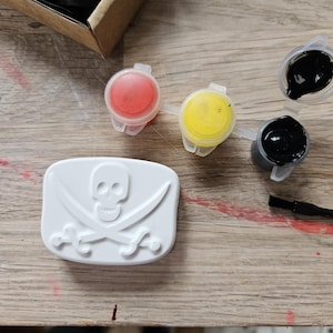 May include: A white rectangular plaster plaque featuring a raised skull and crossbones design. Included are three small plastic pots containing red, yellow, and black paint, along with a thin black paintbrush for applying the paint to the plaque.