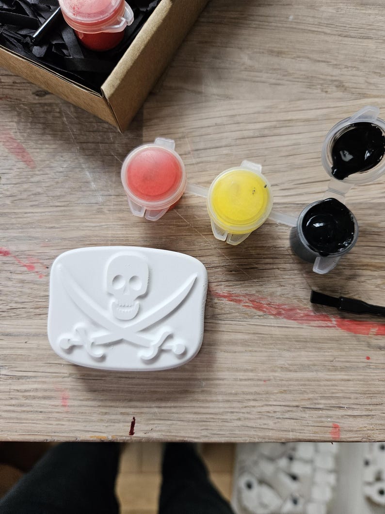 May include: A white rectangular plaster plaque featuring a raised skull and crossbones design. Included are three small plastic pots containing red, yellow, and black paint, along with a thin black paintbrush for applying the paint to the plaque.