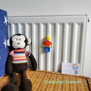 May include: A brown and white striped monkey plush toy sits on a wicker surface next to a white radiator. A yellow, red, and blue Lego figurine is attached to the radiator.