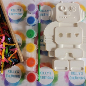 May include: A white ceramic robot figurine with paint pots and a paintbrush. The robot has a round head with two large eyes and a small mouth. The robot's body has a rectangular torso with three circles and a triangle. The robot has two arms and two legs. The paint pots are in various colors, including purple, blue, green, yellow, orange, and red. The paintbrush is green.