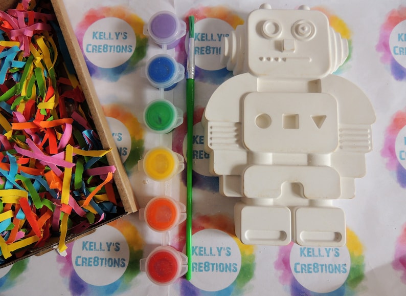 May include: A white ceramic robot figurine with paint pots and a paintbrush. The robot has a round head with two large eyes and a small mouth. The robot's body has a rectangular torso with three circles and a triangle. The robot has two arms and two legs. The paint pots are in various colors, including purple, blue, green, yellow, orange, and red. The paintbrush is green.