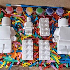 May include: A craft kit with white ceramic figurines of a person and building blocks, along with five small paint pots in red, blue, yellow, green, and purple.