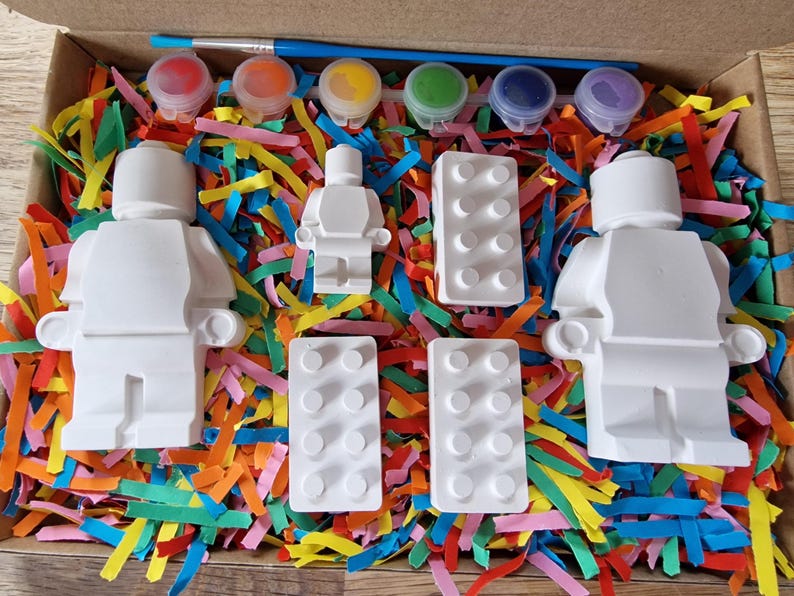 May include: A craft kit with white ceramic figurines of a person and building blocks, along with five small paint pots in red, blue, yellow, green, and purple.