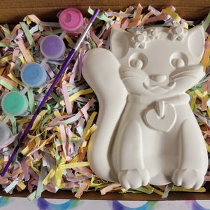Pu&ograve; includere: Una statuetta di gatto in ceramica bianca con un cuore sul petto e fiori sulla testa, in una scatola con un pennello viola e cinque piccoli barattoli di vernice di vari colori.