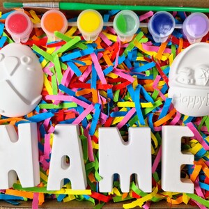 May include: A craft kit with white ceramic letters spelling "NAME", two small ceramic Easter-themed figurines, and four paint pots with colorful paint. The kit is surrounded by colorful shredded paper.