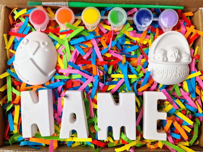 May include: A craft kit with white ceramic letters spelling "NAME", two small ceramic Easter-themed figurines, and four paint pots with colorful paint. The kit is surrounded by colorful shredded paper.
