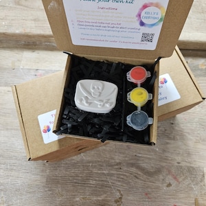 May include: A brown cardboard box containing a paint your own kit with a white skull and crossbones plaster figure, three small paint pots in red, yellow, and black, and a black paintbrush nestled in black paper shred filler material.